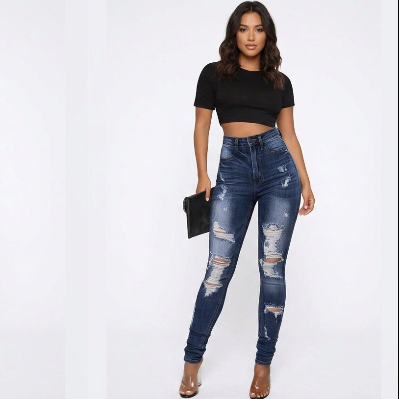 A woman modeling a pair of dark blue high-waisted skinny jeans with distressed rips on the thighs and knees, paired with a black crop top and holding a black clutch against a white studio background.
