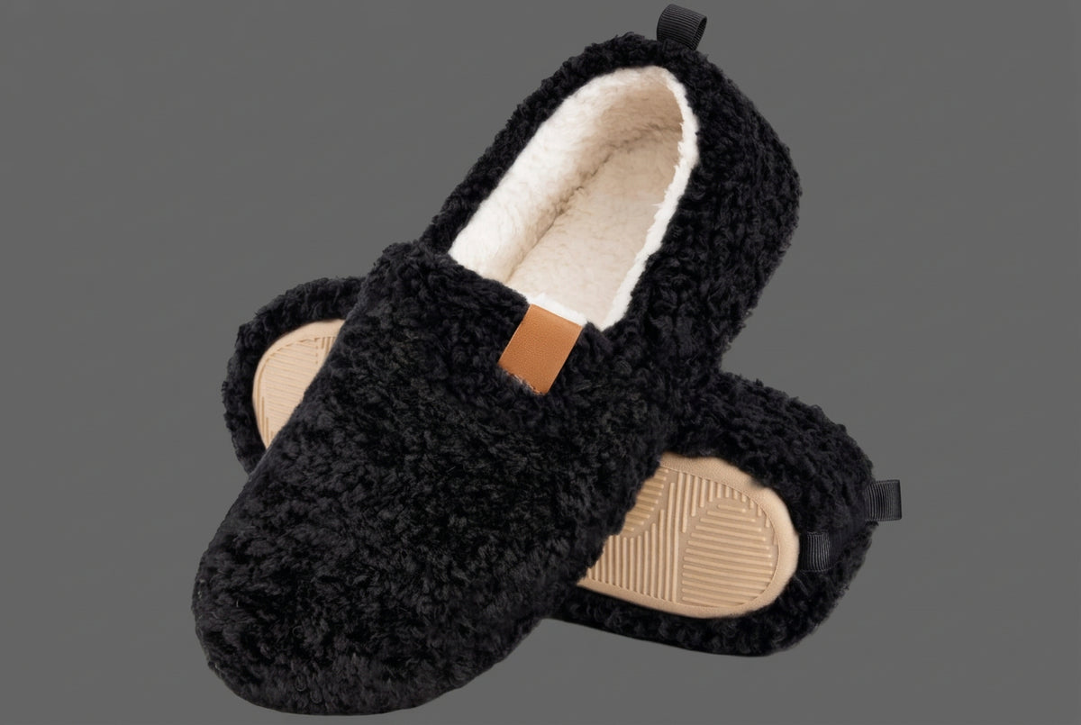 A pair of black sherpa fleece slip-on slippers with a cream lining and a tan outsole, shown on a gray background.