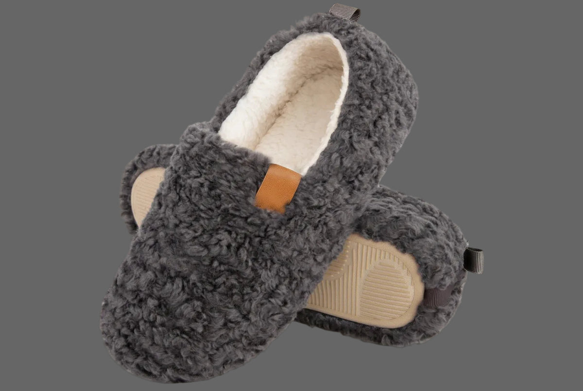 A pair of fuzzy dark gray slip-on slippers with a cream sherpa lining and a small tan accent tab, shown on a solid gray background.