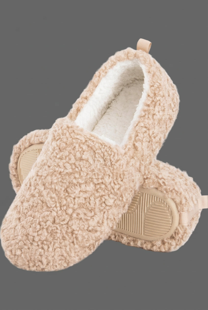 A pair of beige sherpa fleece slip-on slippers with a soft white lining and a small brown leather brand tab on the side, shown on a clean gray background.
