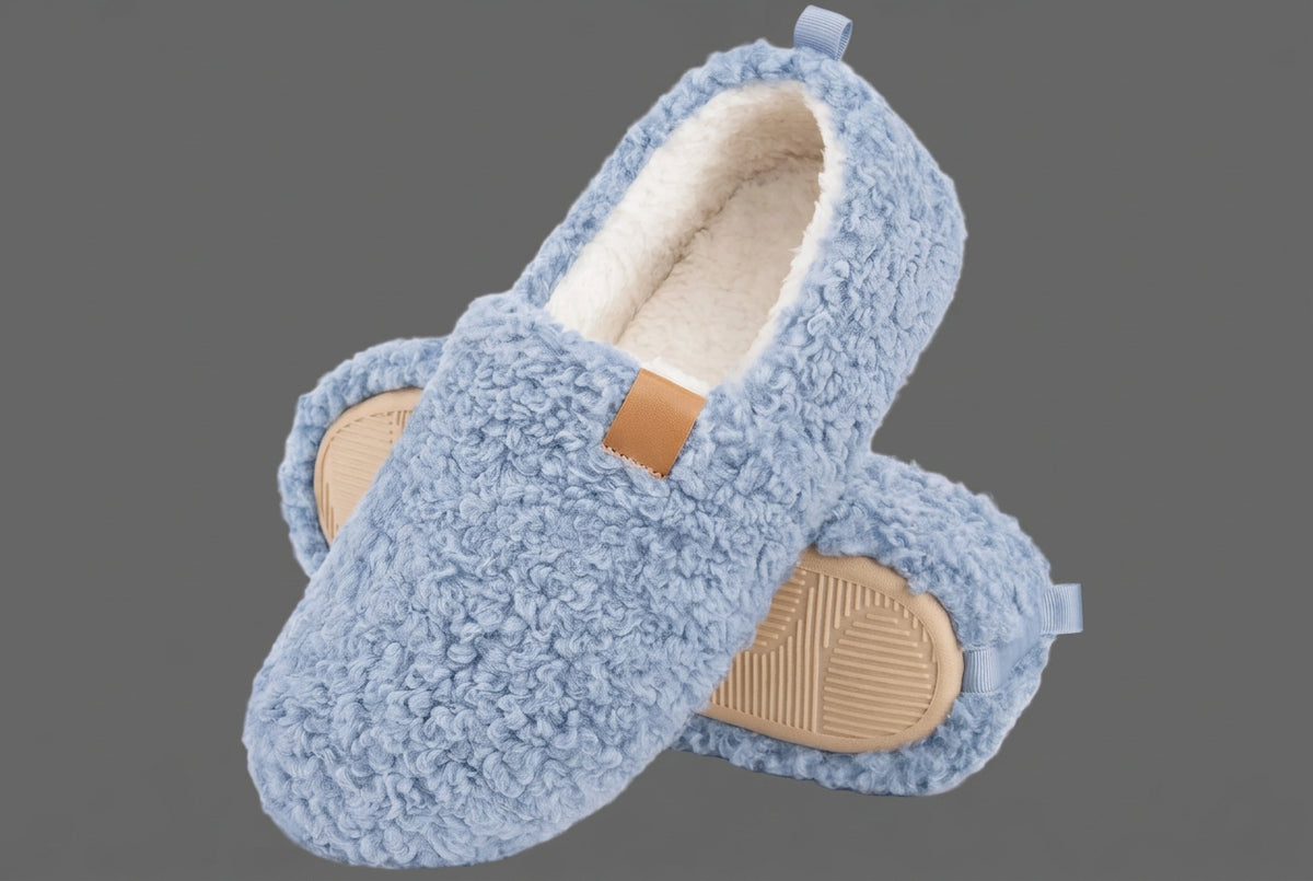 A pair of light blue sherpa fleece slip-on slippers with beige outsoles and a small tan brand patch on the side, shown on a gray background.