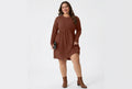 A woman wearing a rust-brown ribbed knit long-sleeve dress with a gathered waist and side pockets, holding a black clutch against a plain white background.
