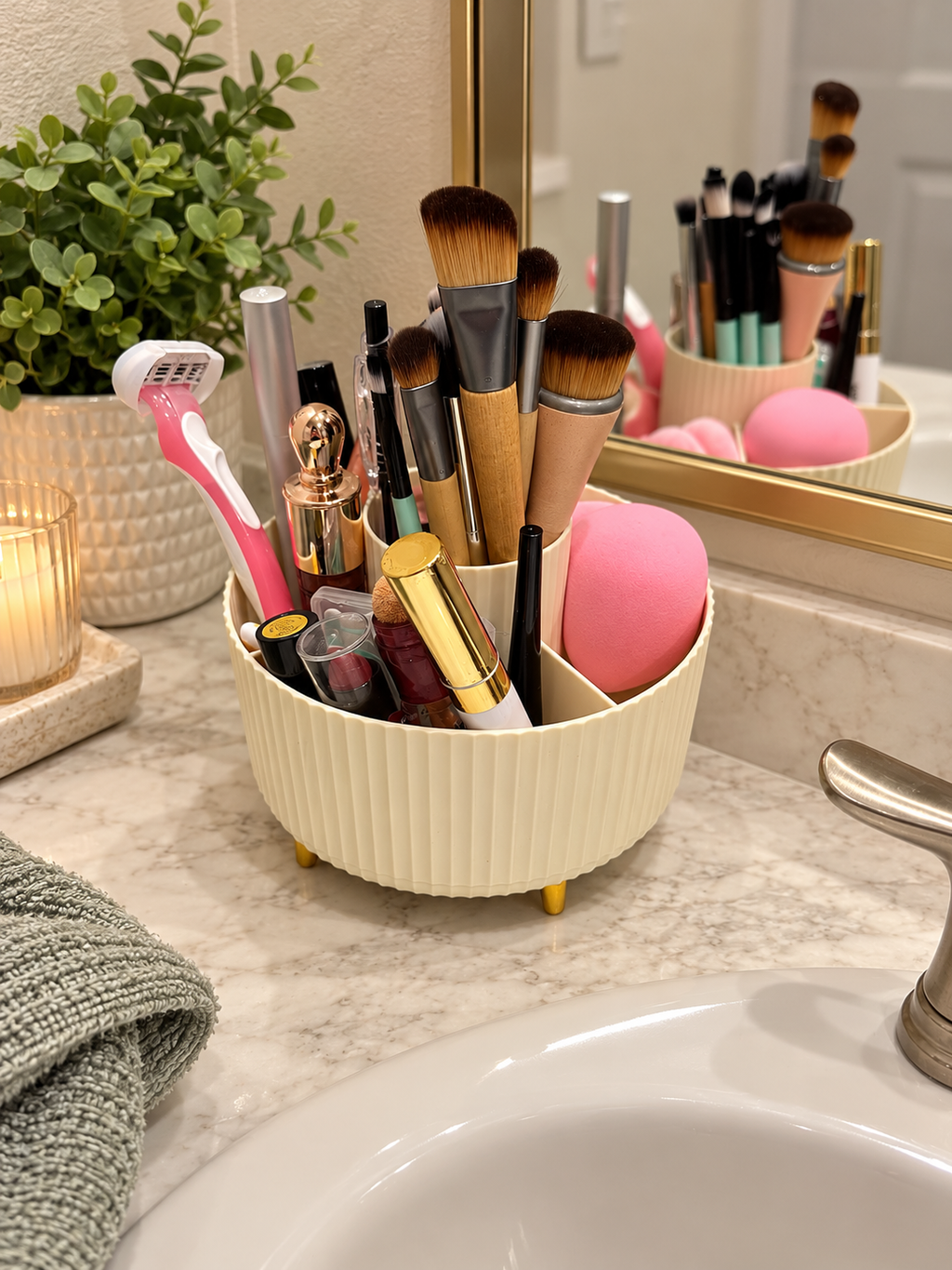 A cream-colored, ribbed round makeup organizer with small gold feet sits on a marble bathroom counter next to a sink. The organizer is filled with makeup brushes, a pink razor, lip gloss, and a pink makeup sponge, with a mirror and bathroom decor visible in the background.