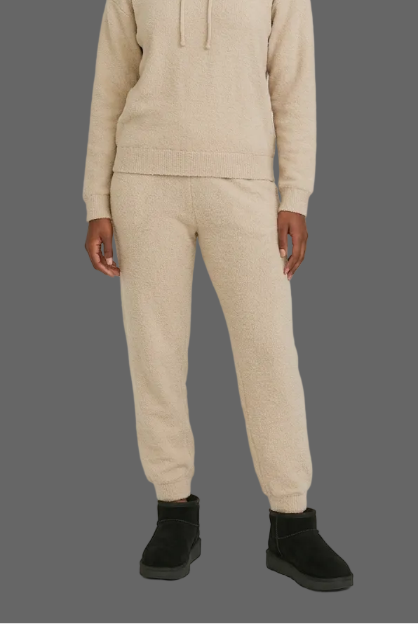 A woman sits cross-legged on a light gray studio floor wearing a beige knit hoodie and jogger set with black ankle boots.